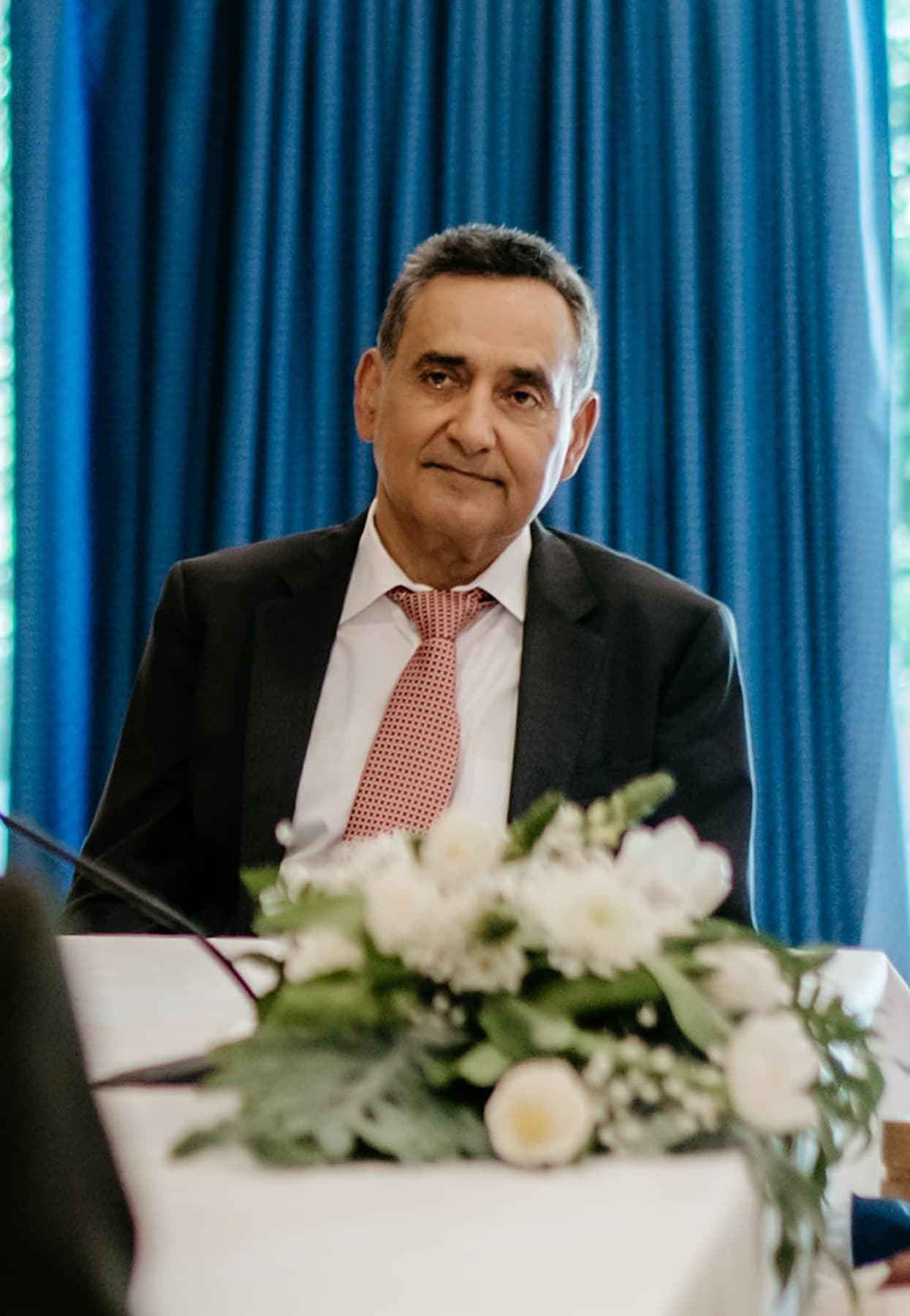Amin Jivraj seated at a white-clothed table before a deep blue drape, half-length, white flowers in the foreground.